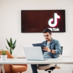 Man using a laptop and tablet in an office setting with a TikTok logo displayed on a wall, illustrating social media engagement strategies.